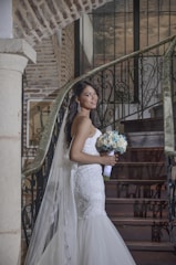A bride in a white lace wedding dress stands on a staircase, holding a bouquet of white and blue flowers. The setting features rustic brick walls and wrought iron railings. Her expression is serene, and she wears elegant earrings.