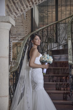 A bride in a white lace wedding dress stands on a staircase, holding a bouquet of white and blue flowers. The setting features rustic brick walls and wrought iron railings. Her expression is serene, and she wears elegant earrings.