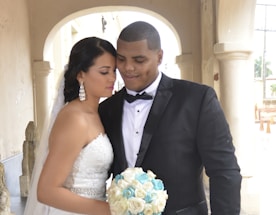 A couple is standing closely under an archway in an elegant setting. The woman is wearing a white strapless wedding dress with intricate lace detailing and holding a bouquet of white and blue flowers. Her long dark hair is styled in a braid. The man is dressed in a black tuxedo with a bow tie, gently leaning his head towards the woman. Both appear serene and content, creating an intimate and loving atmosphere.