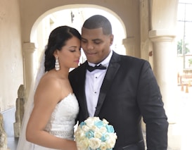 A couple is standing closely under an archway in an elegant setting. The woman is wearing a white strapless wedding dress with intricate lace detailing and holding a bouquet of white and blue flowers. Her long dark hair is styled in a braid. The man is dressed in a black tuxedo with a bow tie, gently leaning his head towards the woman. Both appear serene and content, creating an intimate and loving atmosphere.