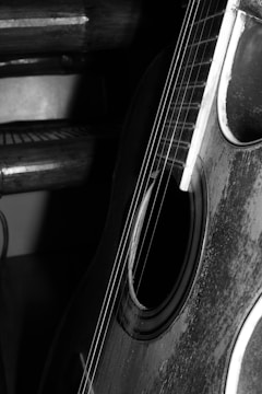 A close-up of an acoustic guitar focusing on the soundhole and strings. The guitar appears aged with visible wear on the body, and the image is in black and white, highlighting the texture and grain of the wood. The background features wooden slats, possibly a chair or bench.