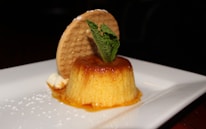A beautifully presented dessert consisting of a creamy flan topped with caramel sauce sits on a white plate. A round biscuit is placed vertically behind the flan, and a sprig of fresh mint is used as garnish. The caramel sauce has elegantly dripped down the sides, and there are small decorative dots of powdered sugar on the plate.