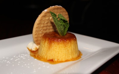 A beautifully presented dessert consisting of a creamy flan topped with caramel sauce sits on a white plate. A round biscuit is placed vertically behind the flan, and a sprig of fresh mint is used as garnish. The caramel sauce has elegantly dripped down the sides, and there are small decorative dots of powdered sugar on the plate.