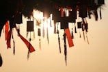 Silhouetted rectangular charms and red tassels hang from a structure, illuminated by the soft glow of a setting sun in the background. The scene suggests tranquility and a sense of reverence.