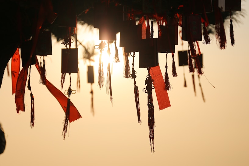 Silhouetted rectangular charms and red tassels hang from a structure, illuminated by the soft glow of a setting sun in the background. The scene suggests tranquility and a sense of reverence.