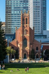A historic red-brick church stands prominently with intricate architectural details, featuring tall arched windows and a central tower. It is set against a backdrop of modern glass skyscrapers. In the foreground, a green lawn is filled with people sitting, walking, and enjoying the sunny day.