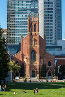 A historic red-brick church stands prominently with intricate architectural details, featuring tall arched windows and a central tower. It is set against a backdrop of modern glass skyscrapers. In the foreground, a green lawn is filled with people sitting, walking, and enjoying the sunny day.
