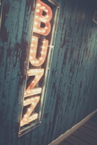 A vertically oriented, illuminated sign spelling out the word 'BUZZ' is mounted on a textured wooden wall with a blue finish. The letters of the sign are large, three-dimensional, and surrounded by glowing light bulbs, creating a warm and inviting glow. The wall features a rustic, weathered appearance with streaks of exposed wood, enhancing the vintage aesthetic.