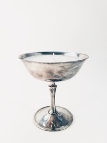 A silver chalice-like object with a single candle wick at its center. The surface is reflective and metallic, suggesting it is made of metal. The background is plain and neutral, enhancing the focus on the chalice.