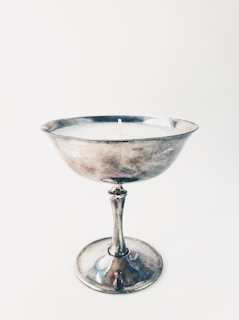 A silver chalice-like object with a single candle wick at its center. The surface is reflective and metallic, suggesting it is made of metal. The background is plain and neutral, enhancing the focus on the chalice.