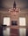 An elegant, spacious room with a large ornate chandelier hanging from the center of a decorated ceiling. Two tall windows with intricate trims allow light to flood the polished wooden floor, creating a reflective surface. A grand piano sits to the side, and a large framed painting hangs between the windows.