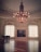 An elegant, spacious room with a large ornate chandelier hanging from the center of a decorated ceiling. Two tall windows with intricate trims allow light to flood the polished wooden floor, creating a reflective surface. A grand piano sits to the side, and a large framed painting hangs between the windows.