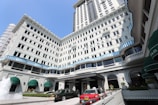 A grand multi-story hotel with white facade and blue striped accents along the windows. Several green awnings adorn the lower part of the building. Luxury cars, including a red taxi, are parked in front of the entrance. A fountain with water jetting upward is visible in the foreground.