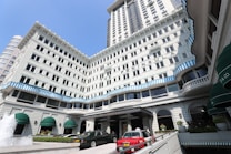 A grand multi-story hotel with white facade and blue striped accents along the windows. Several green awnings adorn the lower part of the building. Luxury cars, including a red taxi, are parked in front of the entrance. A fountain with water jetting upward is visible in the foreground.