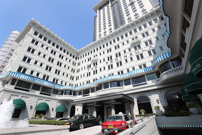A grand multi-story hotel with white facade and blue striped accents along the windows. Several green awnings adorn the lower part of the building. Luxury cars, including a red taxi, are parked in front of the entrance. A fountain with water jetting upward is visible in the foreground.