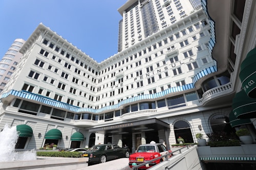 A grand multi-story hotel with white facade and blue striped accents along the windows. Several green awnings adorn the lower part of the building. Luxury cars, including a red taxi, are parked in front of the entrance. A fountain with water jetting upward is visible in the foreground.