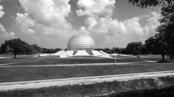A large geodesic dome structure is situated in an open landscape with expansive fields and trees. The sky is filled with scattered clouds, and the overall setting appears tranquil and spacious.