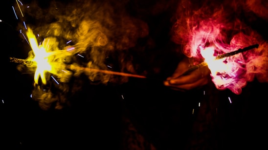 Two sparklers are held by a person, creating vibrant displays of light and smoke against a dark background. One sparkler emits a bright yellow glow while the other produces a vivid red hue. Smoke swirls around the sparks, adding dynamic movement to the scene.