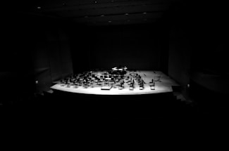 A dimly lit concert hall stage features an arrangement of empty chairs and music stands, creating a sense of stillness and anticipation. The spotlight highlights the stage, contrasting sharply with the darkness of the surrounding auditorium.