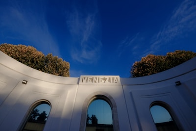A curved white building with tall windows stands under a clear, deep blue sky. The top of the structure displays the word 'Venezia' in bold lettering. Surrounding the top of the building, lush green and brown trees add a touch of nature to the urban architecture.