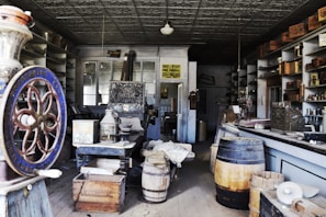 An old-fashioned general store interior featuring vintage equipment and wooden barrels. The room is filled with shelves holding various goods and boxes. A large wheel is prominently displayed in the foreground.