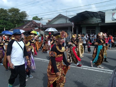 A lively street parade with a diverse group of participants in colorful traditional costumes. Several people, including children, are adorned with intricate headpieces and vibrant fabric, walking along a road lined with onlookers. Some carry umbrellas for shade. The scene is set against a backdrop of residential buildings and trees under a cloudy sky.