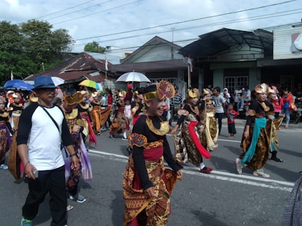 A lively street parade with a diverse group of participants in colorful traditional costumes. Several people, including children, are adorned with intricate headpieces and vibrant fabric, walking along a road lined with onlookers. Some carry umbrellas for shade. The scene is set against a backdrop of residential buildings and trees under a cloudy sky.