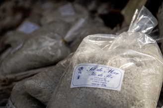 A plastic bag is filled with a coarse white substance, likely sea salt. The bag has a label featuring text and a small drawing. Other similar bags are piled in the background, suggesting a market or store setting.