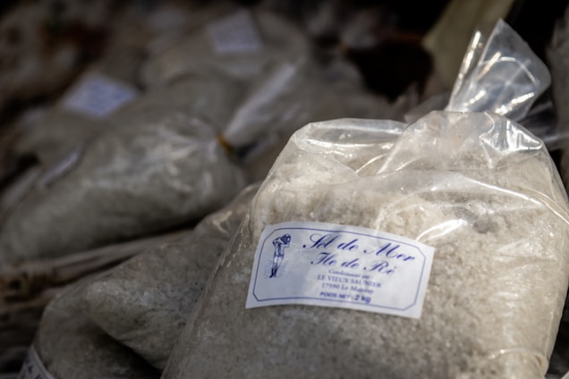 A plastic bag is filled with a coarse white substance, likely sea salt. The bag has a label featuring text and a small drawing. Other similar bags are piled in the background, suggesting a market or store setting.