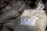 A plastic bag is filled with a coarse white substance, likely sea salt. The bag has a label featuring text and a small drawing. Other similar bags are piled in the background, suggesting a market or store setting.