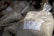 A plastic bag is filled with a coarse white substance, likely sea salt. The bag has a label featuring text and a small drawing. Other similar bags are piled in the background, suggesting a market or store setting.