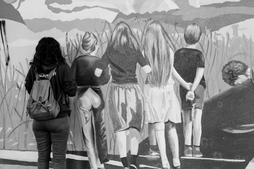 A black and white photograph depicting a person with a backpack standing in front of a wall featuring a mural of four people viewed from behind, looking at a scenic landscape.