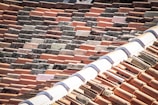 A close-up view of a rooftop covered with variously colored clay tiles. The tiles are arranged in a staggered pattern, showing signs of weathering and age. The roof has a ridge with a lighter, curved tile line running across the top.