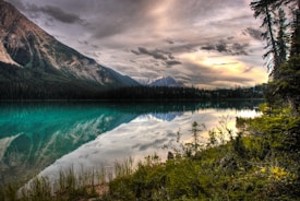 A serene mountain landscape features a clear, turquoise lake reflecting the surrounding tree-lined hills and dramatic cloudy sky. Pine trees frame the scene, while the distant snow-capped mountains enhance the tranquil setting.