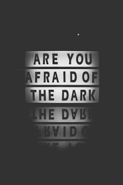 A dark black and white image features a lightbox sign displaying the words 'Are you afraid of the dark' with a reflection of the message below it.