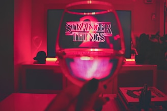 A dimly lit room with a television displaying the title 'Stranger Things 2' in glowing red letters. In front of the screen, there is a wine glass held up, capturing the TV's reflection. The ambience is illuminated with red lighting, creating a cozy and mysterious atmosphere. Books and decorative items are visible on a shelf nearby.