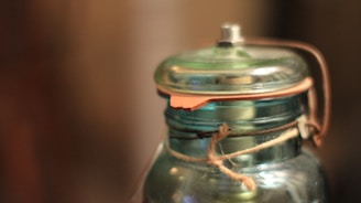A close-up of a transparent glass jar with a metal lid and a leather strap attached. The jar is wrapped with a piece of twine, creating a rustic appearance. The background is blurred, focusing attention on the jar's details and textures.