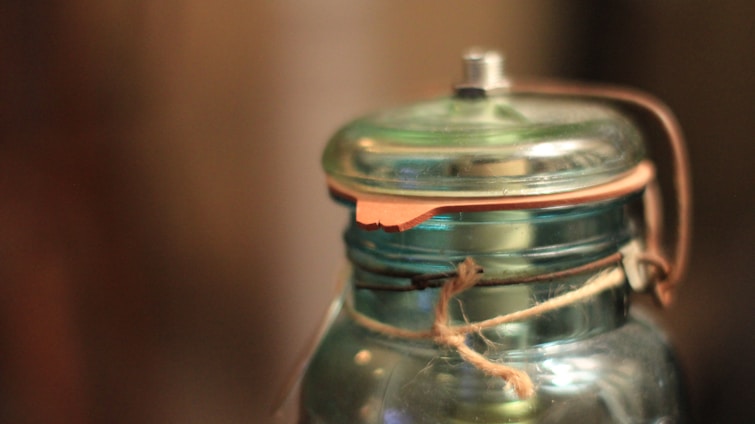 A close-up of a transparent glass jar with a metal lid and a leather strap attached. The jar is wrapped with a piece of twine, creating a rustic appearance. The background is blurred, focusing attention on the jar's details and textures.