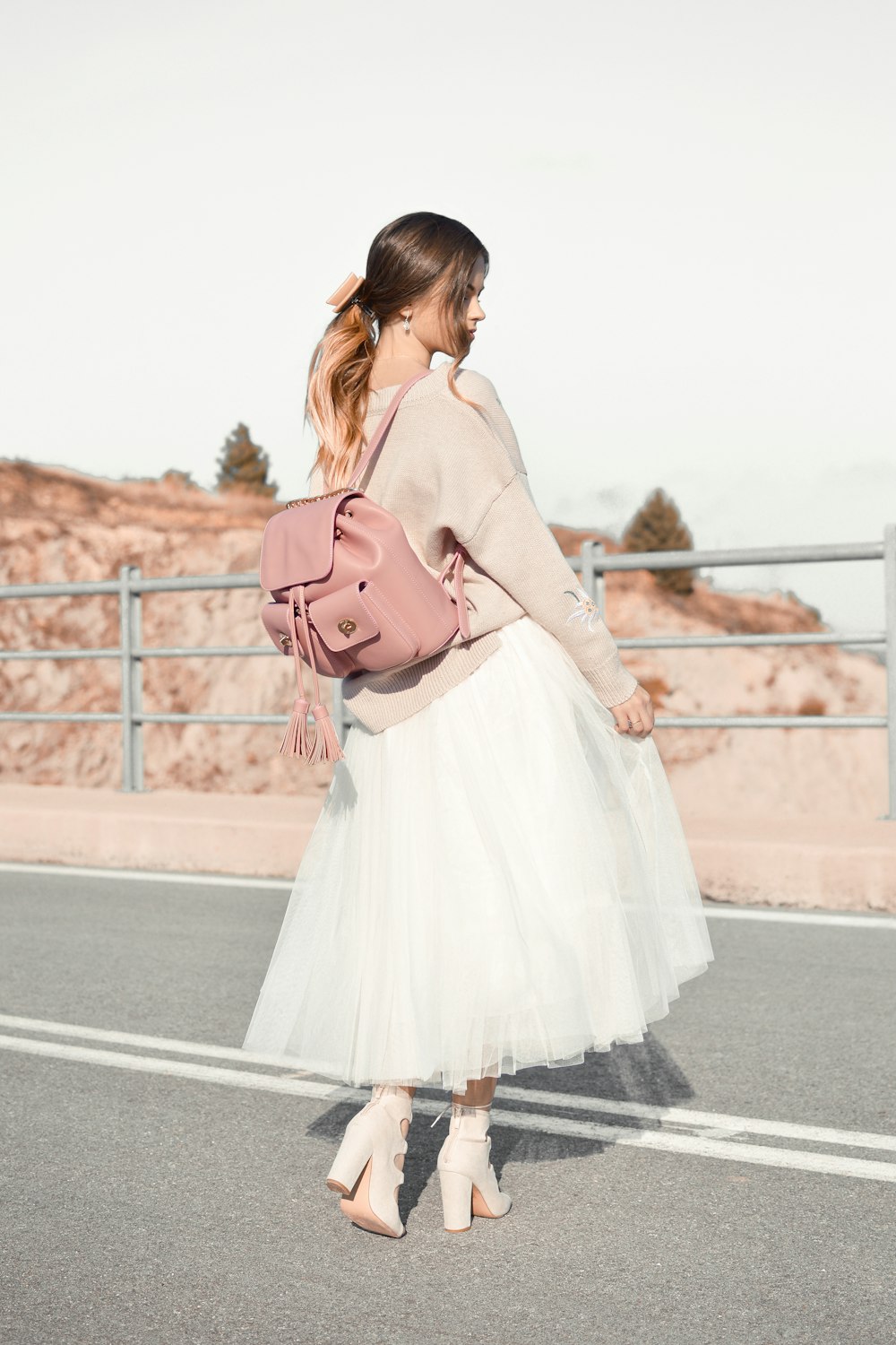 woman standing in front of metal railings wearing brown sweater and white skirt and pair of heeled sandals carrying brown leather backpack during daytime