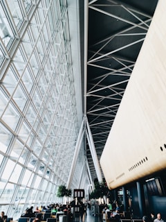 A modern interior space with a high ceiling and glass walls featuring a geometric pattern. There are large structural beams and a wooden paneled area. People are seated in a waiting area, and there is a bar located centrally. Potted plants add greenery to the space.