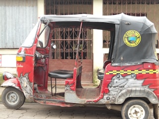 A red and black auto rickshaw with decorative designs on its body is parked on a street. It has an open side with a single seat visible. The hood has an emblem with text, and the background features a building with barred windows and a corrugated metal wall.