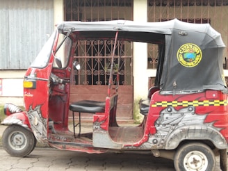 A red and black auto rickshaw with decorative designs on its body is parked on a street. It has an open side with a single seat visible. The hood has an emblem with text, and the background features a building with barred windows and a corrugated metal wall.