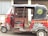 A red and black auto rickshaw with decorative designs on its body is parked on a street. It has an open side with a single seat visible. The hood has an emblem with text, and the background features a building with barred windows and a corrugated metal wall.