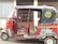 A red and black auto rickshaw with decorative designs on its body is parked on a street. It has an open side with a single seat visible. The hood has an emblem with text, and the background features a building with barred windows and a corrugated metal wall.