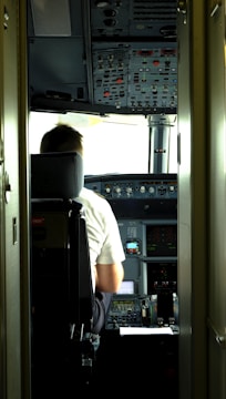 A pilot is seated in the cockpit of an aircraft. The cockpit is filled with numerous control panels, switches, and display screens. Natural light filters in through the windows, illuminating the space.