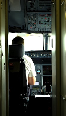 A pilot is seated in the cockpit of an aircraft. The cockpit is filled with numerous control panels, switches, and display screens. Natural light filters in through the windows, illuminating the space.