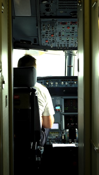 A pilot is seated in the cockpit of an aircraft. The cockpit is filled with numerous control panels, switches, and display screens. Natural light filters in through the windows, illuminating the space.