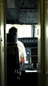 A pilot is seated in the cockpit of an aircraft. The cockpit is filled with numerous control panels, switches, and display screens. Natural light filters in through the windows, illuminating the space.