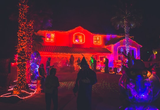A house is adorned with an abundance of bright red and orange lights, creating an eerie, festive atmosphere. Silhouetted figures in Halloween costumes move around the driveway, and various Halloween decorations, including a large inflatable black cat and pumpkins, are scattered around. Two palm trees are wrapped in orange lights, enhancing the spooky theme.