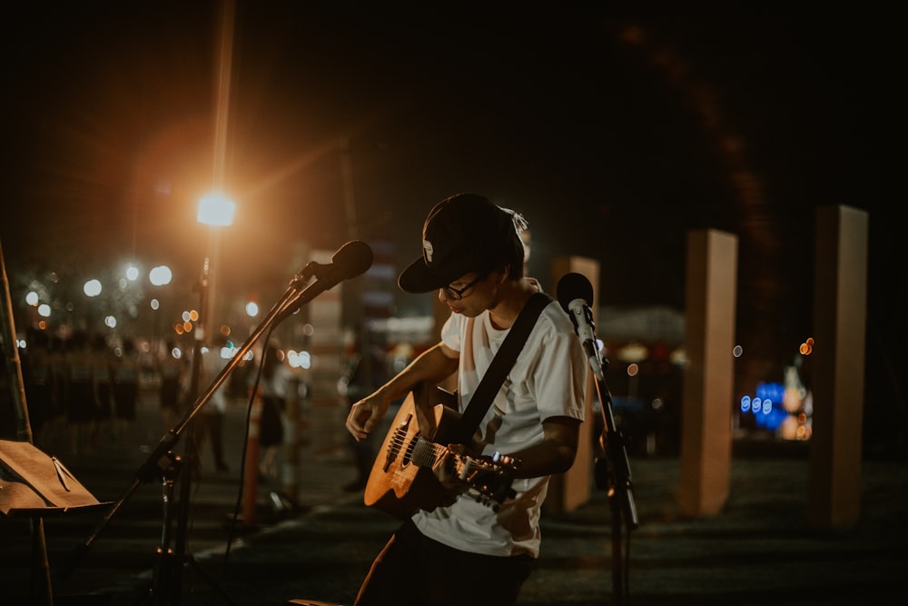 흰색 티셔츠를 입고 기타를 연주하는 남자가 야간에 열린 공간에 스탠드가 있는 마이크 앞에 서 있습니다.